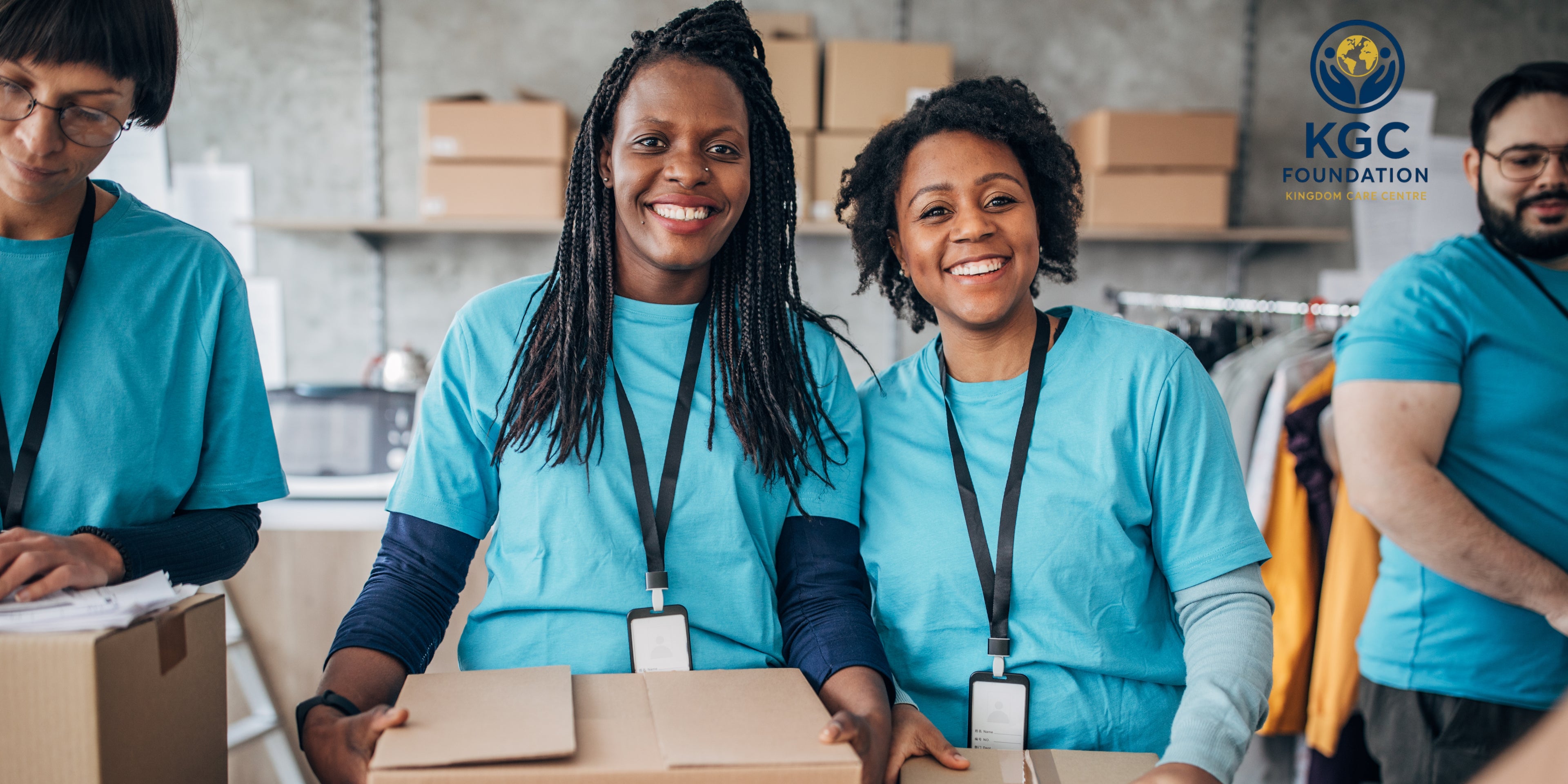 Two women in blue shirts with lanyards standing in a warehouse setting, smiling at the camera. KGC Foundation logo visible.